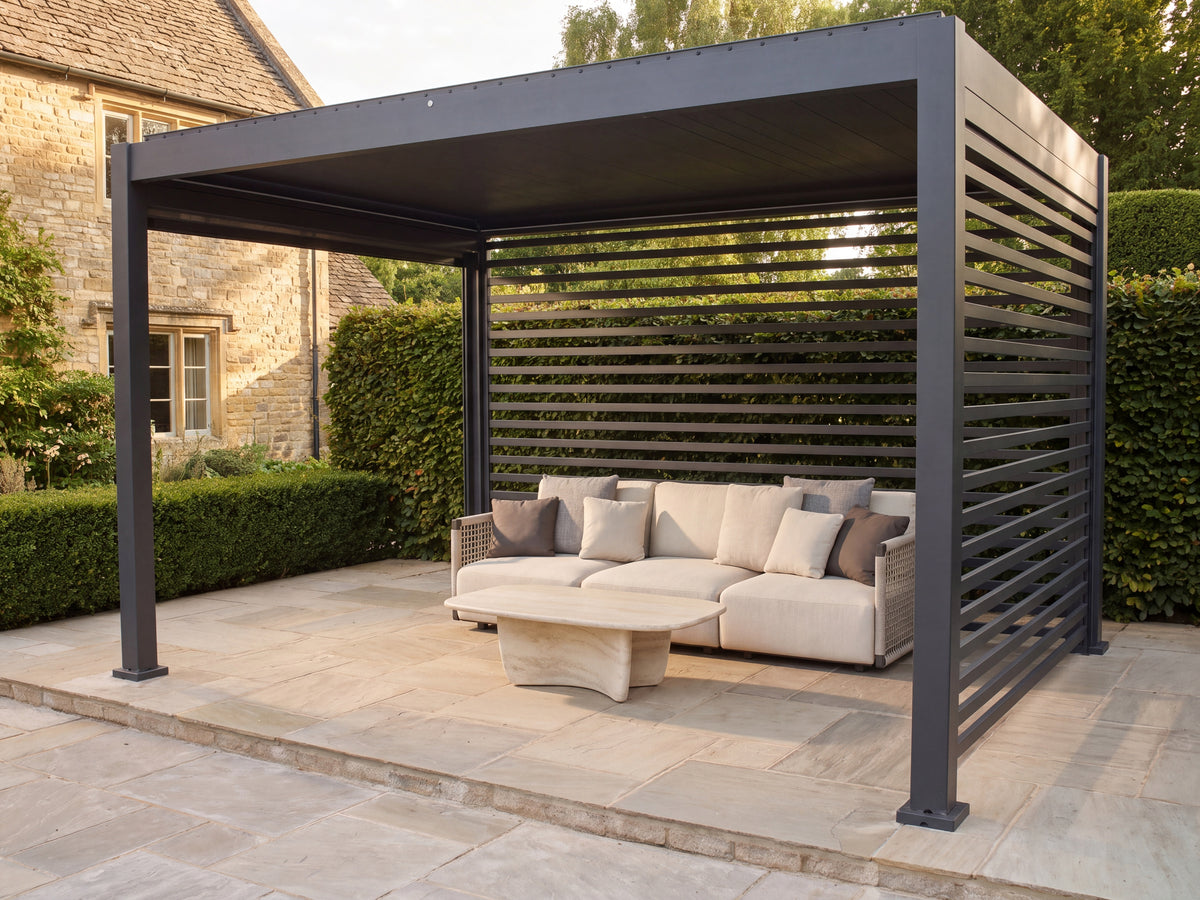 Como pergola with a closed roof and slatted walls over a cream outdoor lounge beside a stone house.
