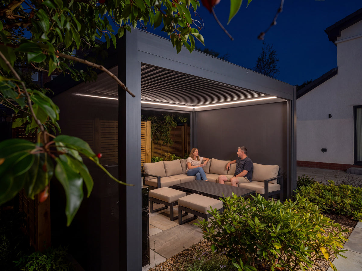 Como pergola at night with open louvres, lowered side screens and white lighting above a couple relaxing in a garden lounge.